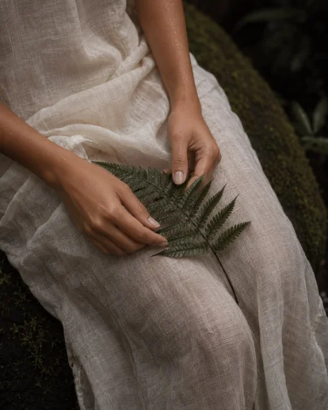 A woman seated in a linen dress, holding a fern frond in her lap